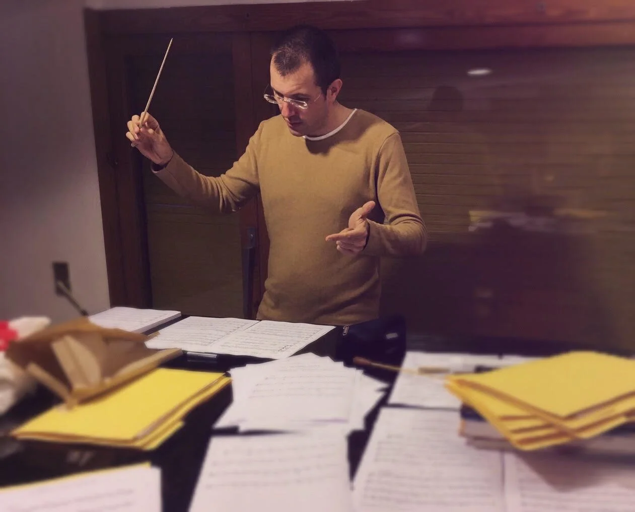 Gerard Pastor rehearsing conducting alone, smiling, with loose sheet music spread out on his piano.
