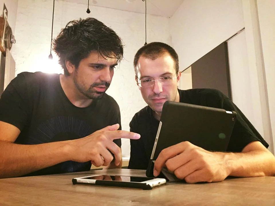 Guillem Barceló and Gerard Pastor, both in black short-sleeve T-shirts, looking at and discussing something on a tablet, seated at a café table.
