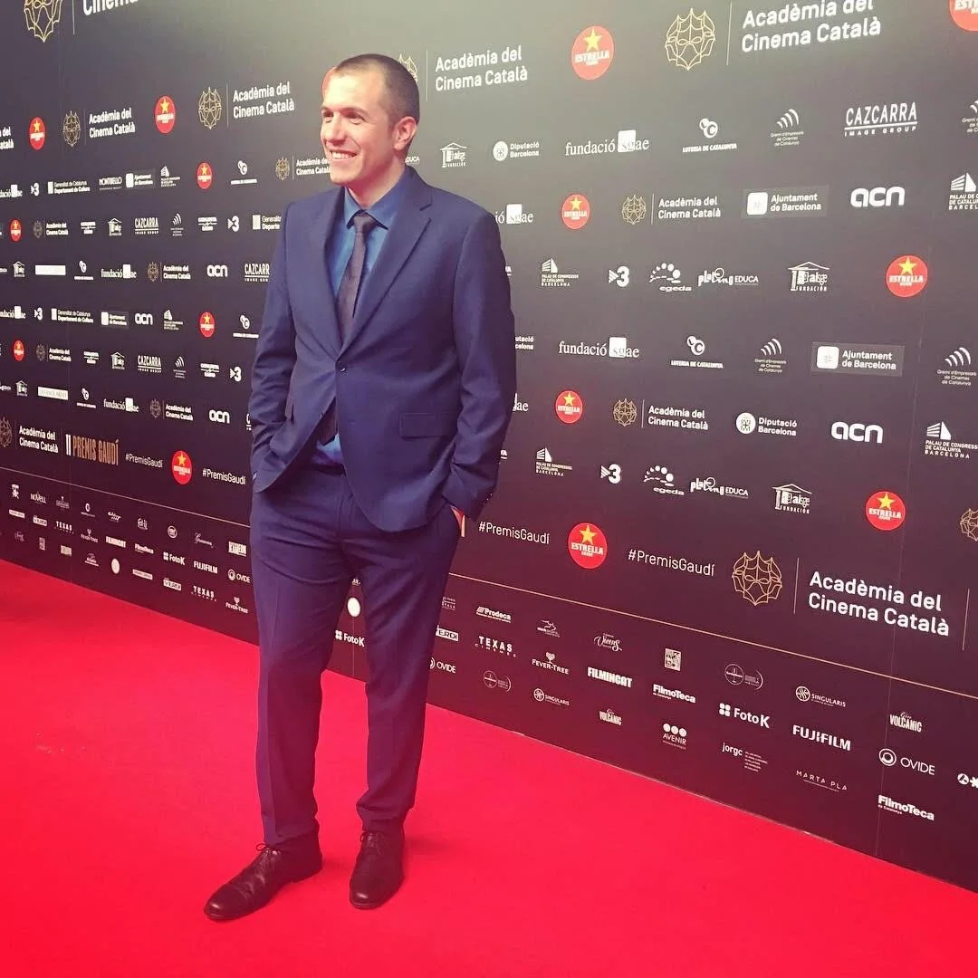 Gerard Pastor in a blue suit and tie on a red carpet, in front of a backdrop with logos and texts in Catalan, smiling with his hands in his pockets. Gaudí Awards Gala.