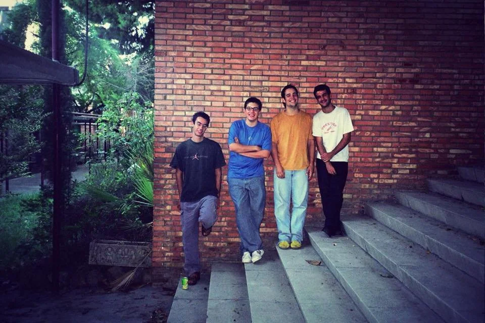 Four young people in casual clothes leaning against a brick wall, looking at the camera.