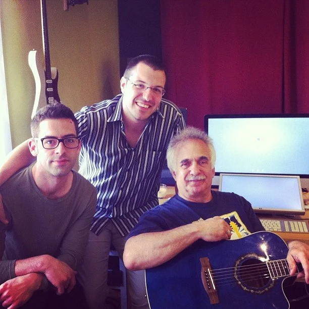 ván Garrido, Jerry Zolten, and Gerard Pastor in a recording studio, all looking at the camera, with Jerry Zolten holding an acoustic guitar.