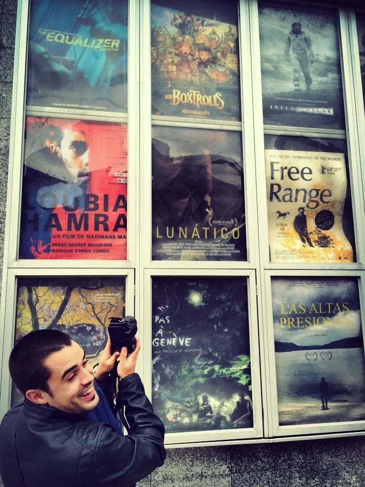 Adri Mena taking a picture of a movie billboard, turning his head toward the camera and smiling.”